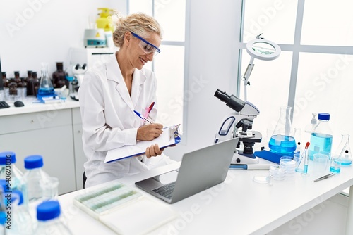 Middle age blonde woman wearing scientist uniform writing on clipboard working at laboratory