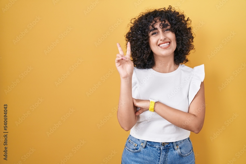 Young middle east woman standing over yellow background smiling with happy face winking at the camera doing victory sign. number two. Young middle east woman standing over yellow background smiling with happy face winking at the camera doing victory sign. number two.