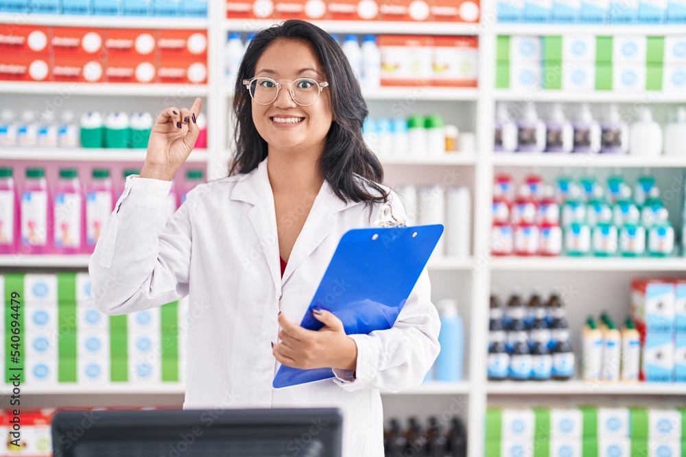 Asian young woman working at pharmacy drugstore holding clipboard surprised with an idea or question pointing finger with happy face, number one