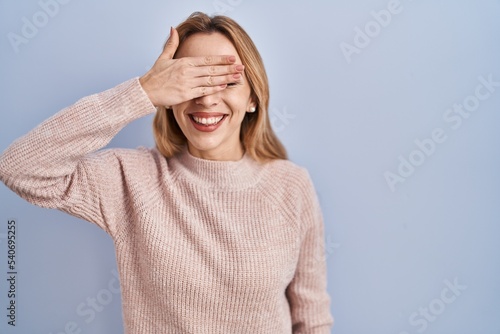 Hispanic woman standing over blue background smiling and laughing with hand on face covering eyes for surprise. blind concept.