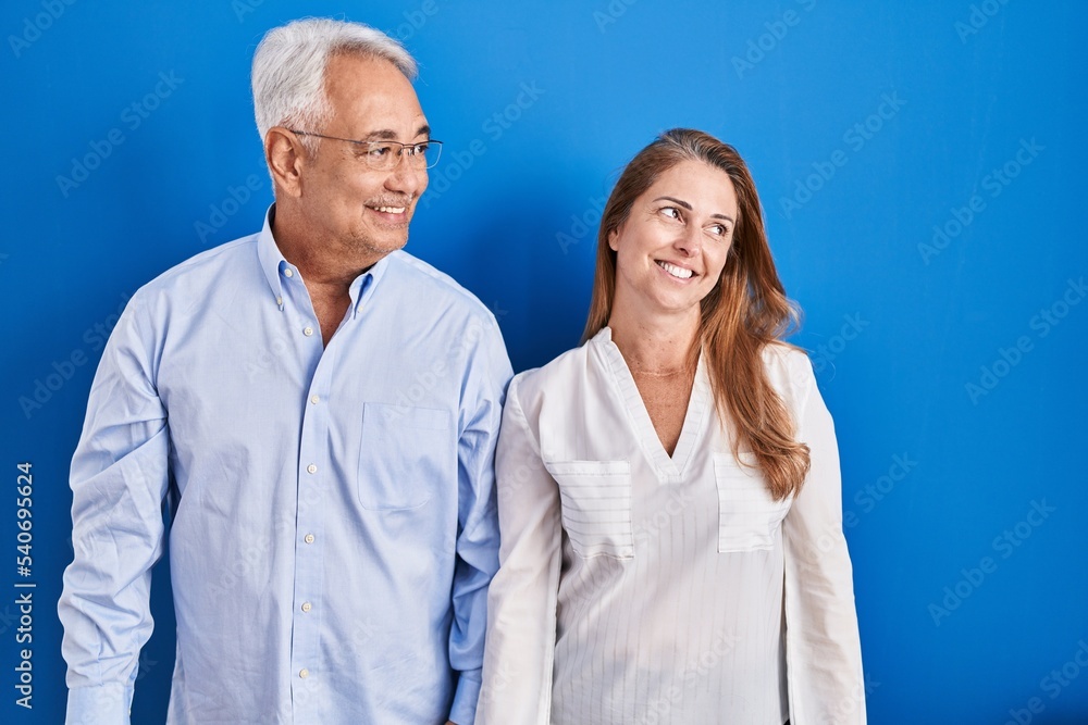 Middle age hispanic couple standing over blue background looking away to side with smile on face, natural expression. laughing confident. Middle age hispanic couple standing over blue background looking away to side with smile on face, natural expression. laughing confident.