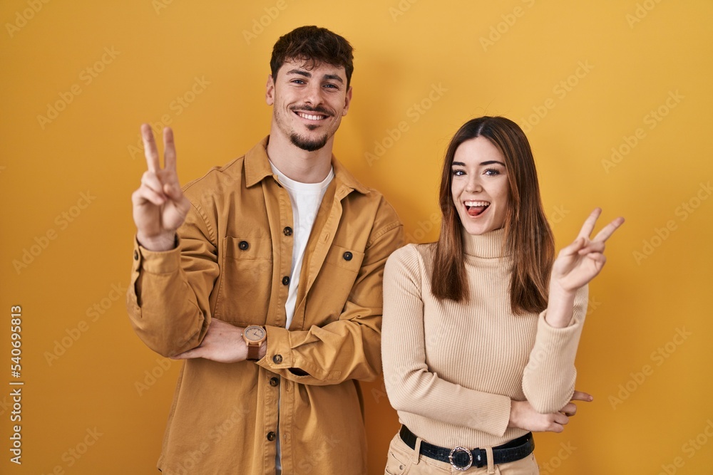 Young hispanic couple standing over yellow background smiling with happy face winking at the camera doing victory sign with fingers. number two. Young hispanic couple standing over yellow background smiling with happy face winking at the camera doing victory sign with fingers. number two.
