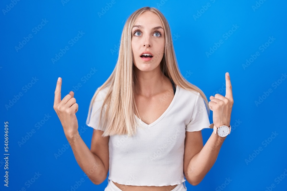 Fototapeta premium Young caucasian woman standing over blue background amazed and surprised looking up and pointing with fingers and raised arms.