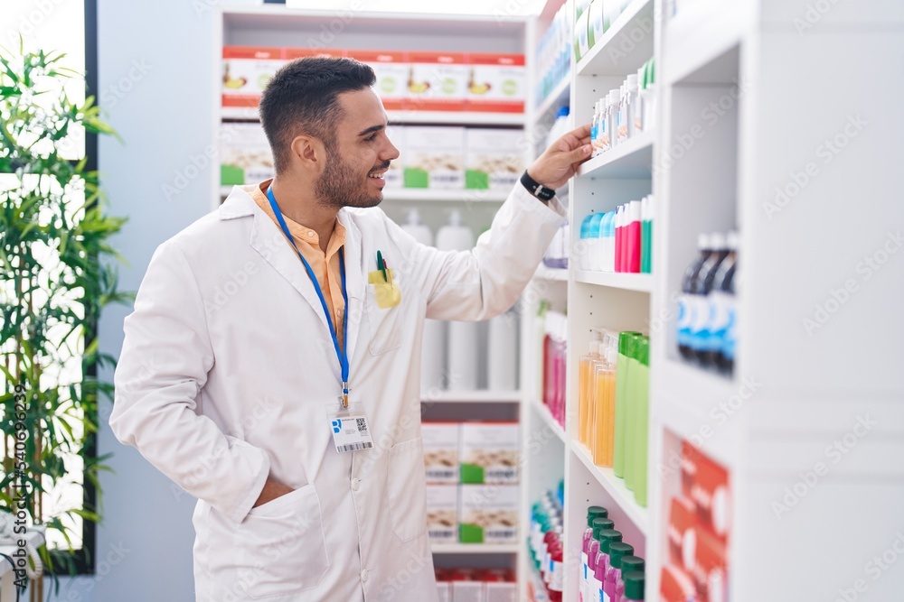 Young hispanic man pharmacist smiling confident holding product on shelving at pharmacy