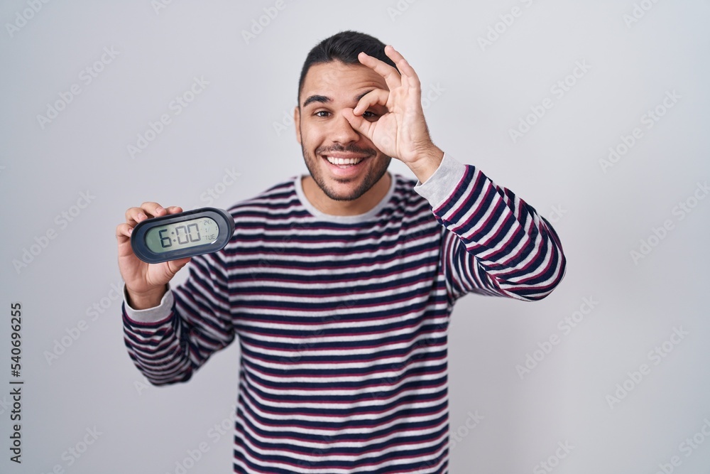 Fototapeta premium Young hispanic man wearing pyjama holding alarm clock smiling happy doing ok sign with hand on eye looking through fingers