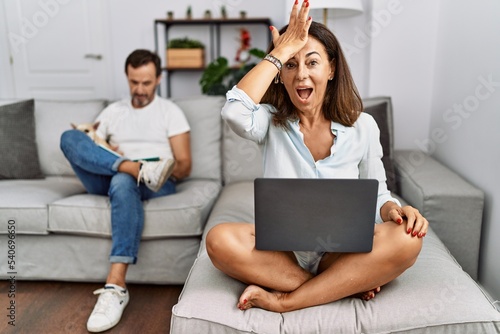 Hispanic middle age couple at home, woman using laptop surprised with hand on head for mistake, remember error. forgot, bad memory concept. © Krakenimages.com