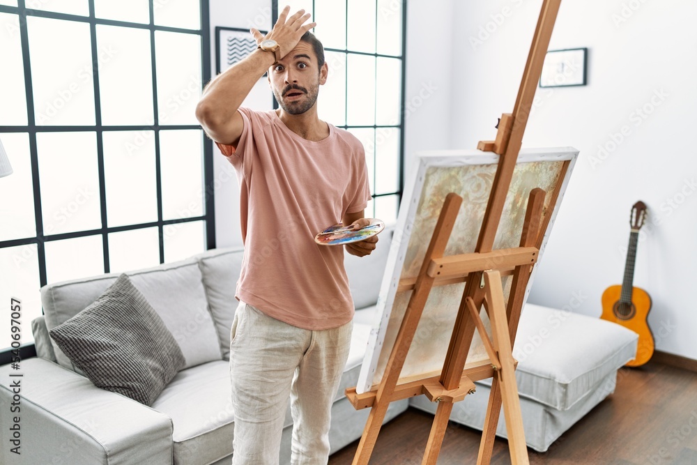Young hispanic man with beard painting on canvas at home surprised with hand on head for mistake, remember error. forgot, bad memory concept.