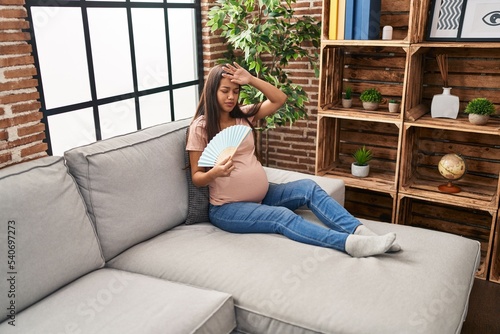 Young latin woman pregnant using hand fan sitting on sofa at home