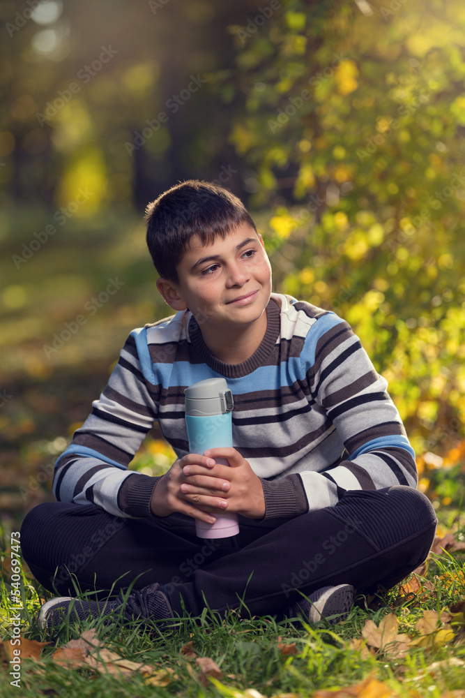 A teenager rests sitting on the grass, in an autumn park, after an active walk, holding a thermos in his hands A teenager rests sitting on the grass, in an autumn park, after an active walk, holding a thermos in his hands