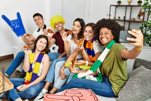 Group of young friends watching and supporting soccer match eating pizza and make selfie by the smartphone at home.