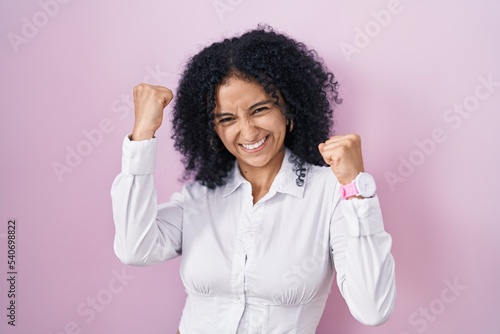 Hispanic woman with curly hair standing over pink background very happy and excited doing winner gesture with arms raised, smiling and screaming for success. celebration concept.