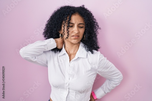 Hispanic woman with curly hair standing over pink background suffering of neck ache injury, touching neck with hand, muscular pain