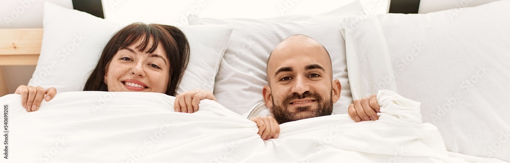 Young hispanic couple covering face with bedsheet lying on the bed at bedroom. Young hispanic couple covering face with bedsheet lying on the bed at bedroom.