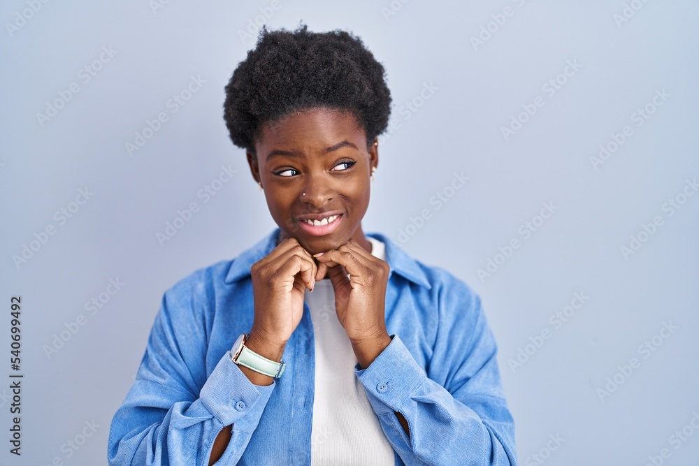 African american woman standing over blue background laughing nervous and excited with hands on chin looking to the side African american woman standing over blue background laughing nervous and excited with hands on chin looking to the side