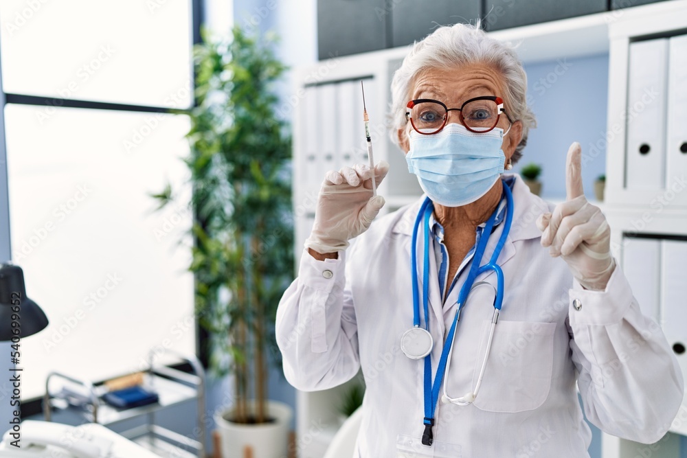 Fototapeta premium Senior woman with grey hair wearing doctor uniform and medical mask holding syringe surprised with an idea or question pointing finger with happy face, number one
