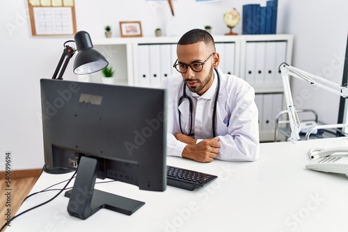 Young hispanic man wearing doctor uniform having video call at clinic