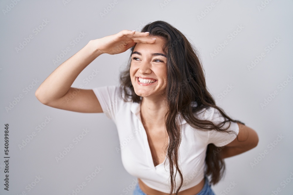Young teenager girl standing over white background very happy and smiling looking far away with hand over head. searching concept. Young teenager girl standing over white background very happy and smiling looking far away with hand over head. searching concept.