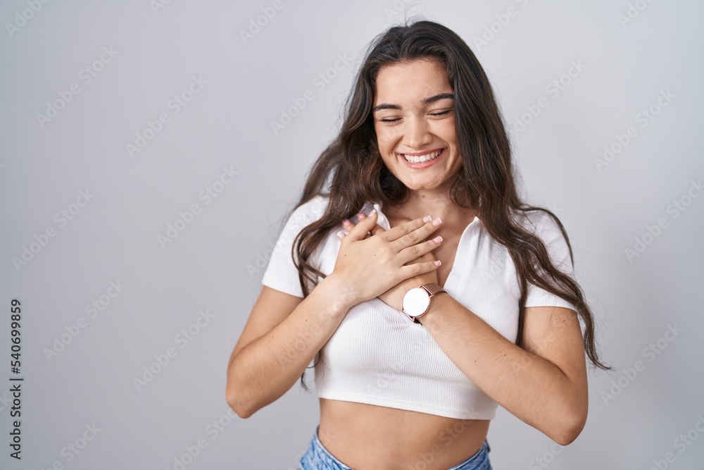 Young teenager girl standing over white background smiling with hands on chest with closed eyes and grateful gesture on face. health concept. Young teenager girl standing over white background smiling with hands on chest with closed eyes and grateful gesture on face. health concept.