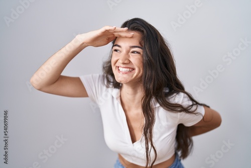 Young teenager girl standing over white background very happy and smiling looking far away with hand over head. searching concept.