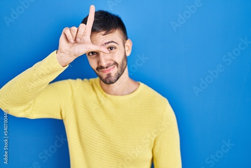 Hispanic man standing over blue background making fun of people with fingers on forehead doing loser gesture mocking and insulting.
