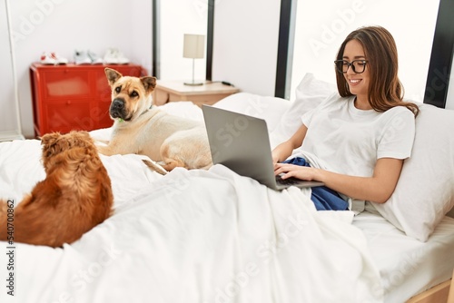 Young hispanic woman using laptop sitting on bed with dogs at bedroom