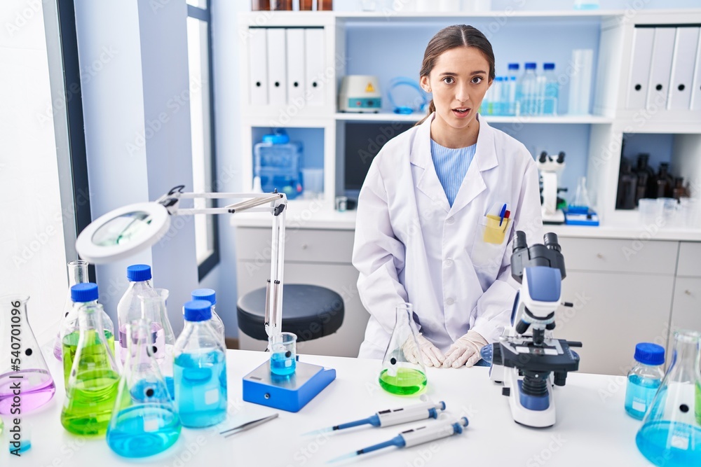Young brunette woman working at scientist laboratory scared and amazed with open mouth for surprise, disbelief face Young brunette woman working at scientist laboratory scared and amazed with open mouth for surprise, disbelief face