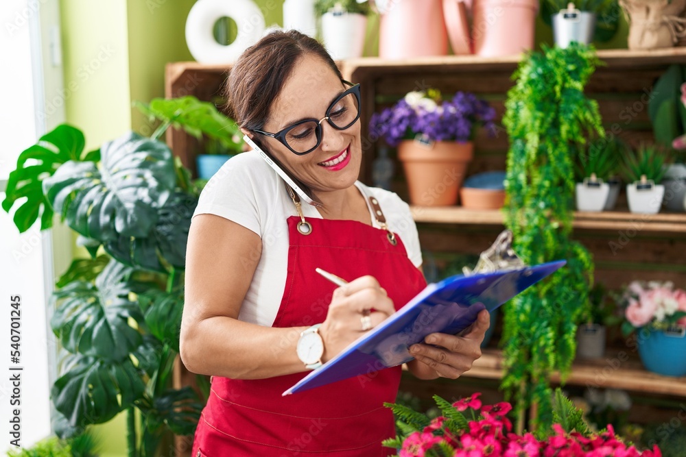 Young beautiful hispanic woman florist talking on smartphone writing on document at flower shop