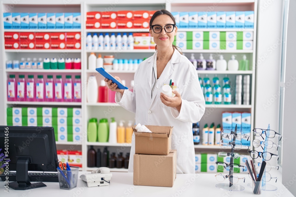 Young beautiful hispanic woman pharmacist using touchpad holding pills bottle at pharmacy