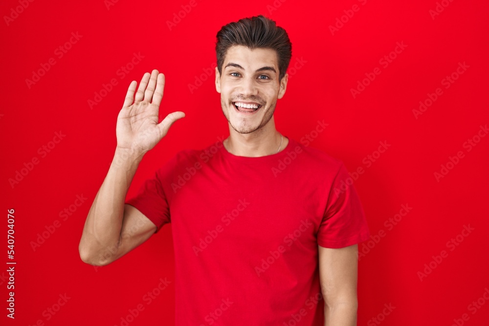Naklejka premium Young hispanic man standing over red background waiving saying hello happy and smiling, friendly welcome gesture