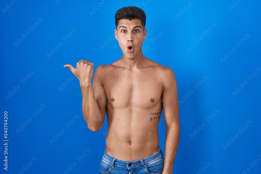Young hispanic man standing shirtless over blue background surprised pointing with hand finger to the side, open mouth amazed expression.