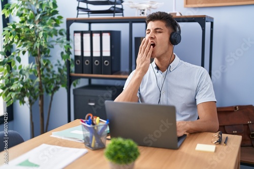 Bilde på lerret Young hispanic man working at the office wearing headphones bored yawning tired covering mouth with hand