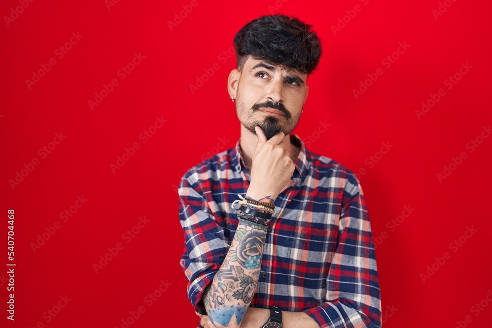Young hispanic man with beard standing over red background with hand on chin thinking about question, pensive expression. smiling with thoughtful face. doubt concept. Young hispanic man with beard standing over red background with hand on chin thinking about question, pensive expression. smiling with thoughtful face. doubt concept.