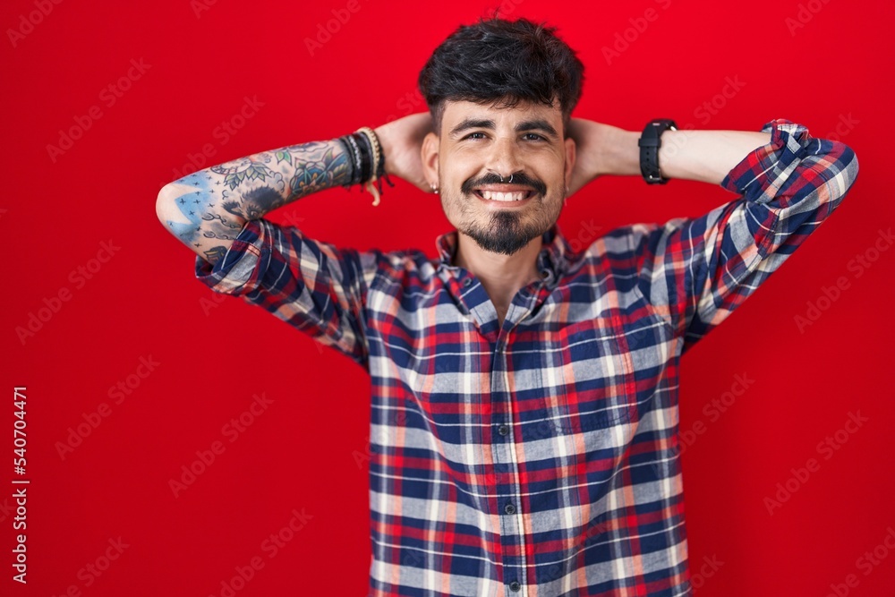 Young hispanic man with beard standing over red background relaxing and stretching, arms and hands behind head and neck smiling happy Young hispanic man with beard standing over red background relaxing and stretching, arms and hands behind head and neck smiling happy
