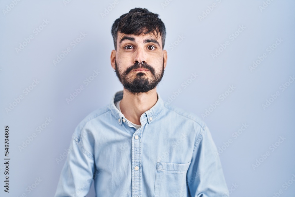 Young hispanic man with beard standing over blue background puffing cheeks with funny face. mouth inflated with air, crazy expression.