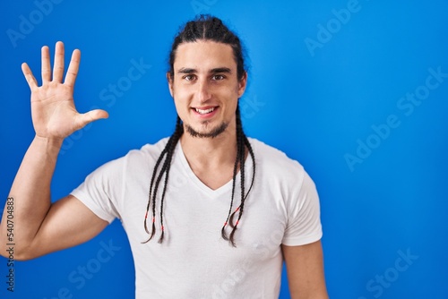 Hispanic man with long hair standing over blue background showing and pointing up with fingers number five while smiling confident and happy.