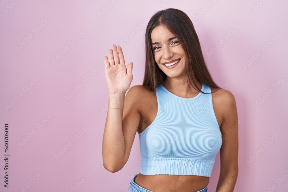 Young brunette woman standing over pink background waiving saying hello happy and smiling, friendly welcome gesture