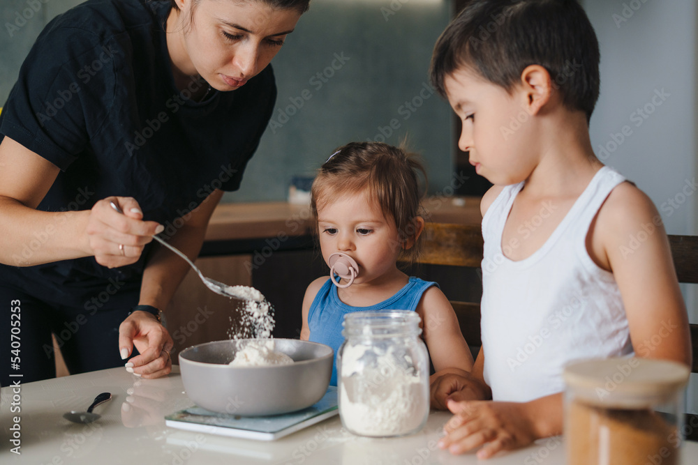 young mother with her two kids weighing out flour into bowl in kitchen environment, baking cakes for the party. Weighing indigrients on electronic kitchen young mother with her two kids weighing out flour into bowl in kitchen environment, baking cakes for the party. Weighing indigrients on electronic kitchen