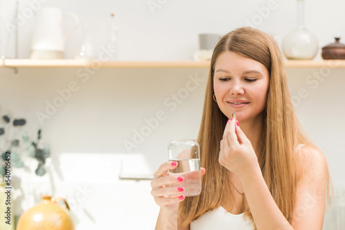 Happy blonde woman holding a glass of water and a capsule of fish oil or vitamin D in the kitchen. Healthy lifestyle.