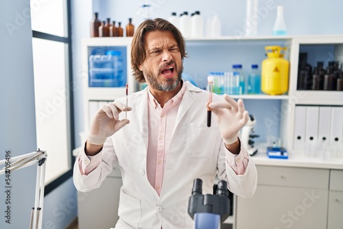 Handsome middle age man holding blood sample at biology laboratory angry and mad screaming frustrated and furious, shouting with anger. rage and aggressive concept.