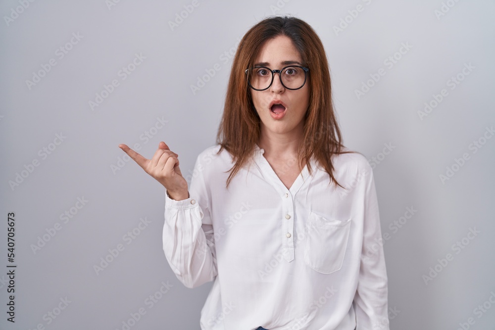 Brunette woman standing over white isolated background surprised pointing with finger to the side, open mouth amazed expression. Brunette woman standing over white isolated background surprised pointing with finger to the side, open mouth amazed expression.
