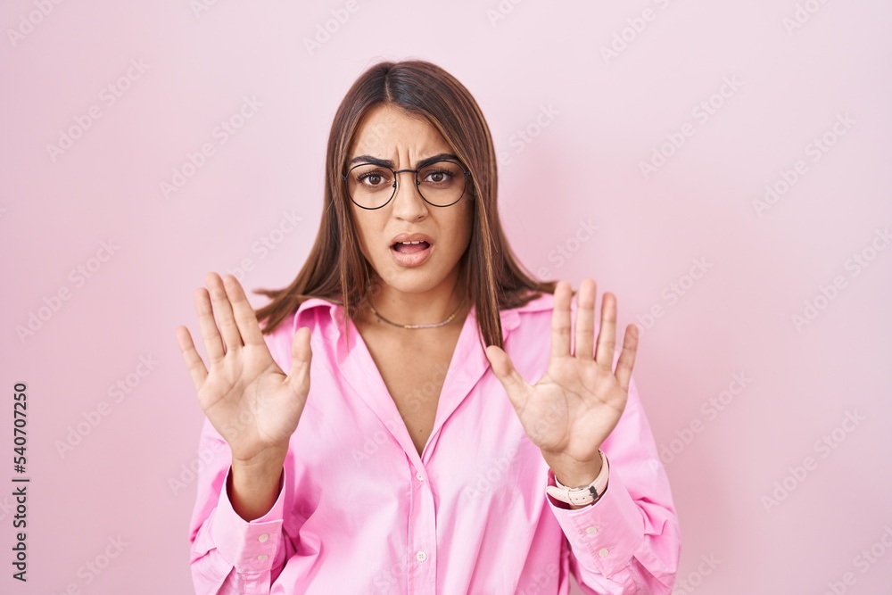 Young hispanic woman wearing glasses standing over pink background moving away hands palms showing refusal and denial with afraid and disgusting expression. stop and forbidden. Young hispanic woman wearing glasses standing over pink background moving away hands palms showing refusal and denial with afraid and disgusting expression. stop and forbidden.
