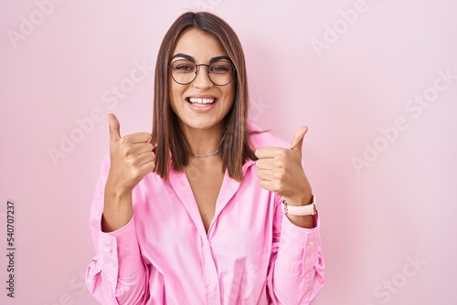 Young hispanic woman wearing glasses standing over pink background success sign doing positive gesture with hand  thumbs up smiling and happy. cheerful expression and winner gesture.