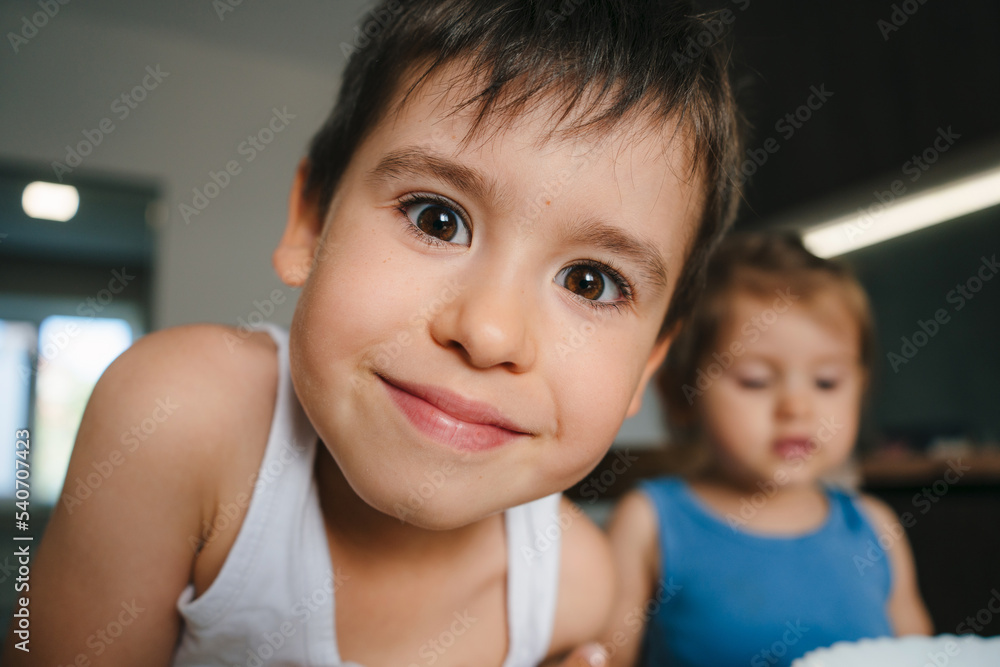 Funny portrait of a boy looking intently at the camera while preparing a cake with his family in the kitchen, weighing the ingredients written in the recipe Funny portrait of a boy looking intently at the camera while preparing a cake with his family in the kitchen, weighing the ingredients written in the recipe