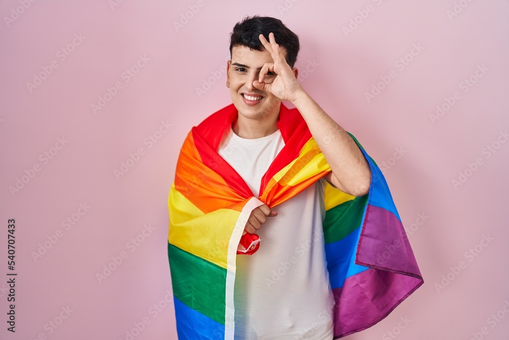 Non binary person holding rainbow lgbtq flag smiling happy doing ok sign with hand on eye looking through fingers Non binary person holding rainbow lgbtq flag smiling happy doing ok sign with hand on eye looking through fingers