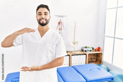 Young handsome man with beard working at pain recovery clinic gesturing with hands showing big and large size sign  measure symbol. smiling looking at the camera. measuring concept.