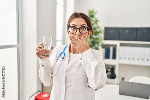 Young brunette doctor woman holding glass of water covering mouth with hand  shocked and afraid for mistake. surprised expression