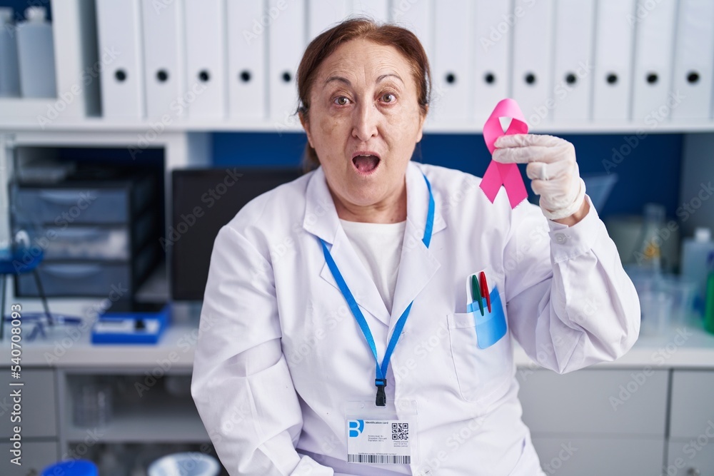 Senior woman with glasses working at scientist laboratory holding pink ribbon scared and amazed with open mouth for surprise, disbelief face Senior woman with glasses working at scientist laboratory holding pink ribbon scared and amazed with open mouth for surprise, disbelief face