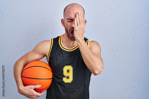 Young bald man with beard wearing basketball uniform holding ball yawning tired covering half face, eye and mouth with hand. face hurts in pain.