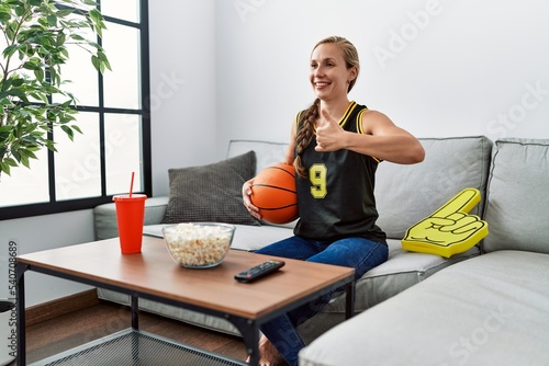 Young blonde woman holding basketball ball cheering tv game smiling happy and positive, thumb up doing excellent and approval sign