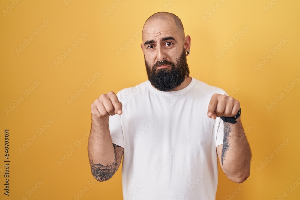 Young hispanic man with beard and tattoos standing over yellow background pointing down looking sad and upset, indicating direction with fingers, unhappy and depressed. Young hispanic man with beard and tattoos standing over yellow background pointing down looking sad and upset, indicating direction with fingers, unhappy and depressed.